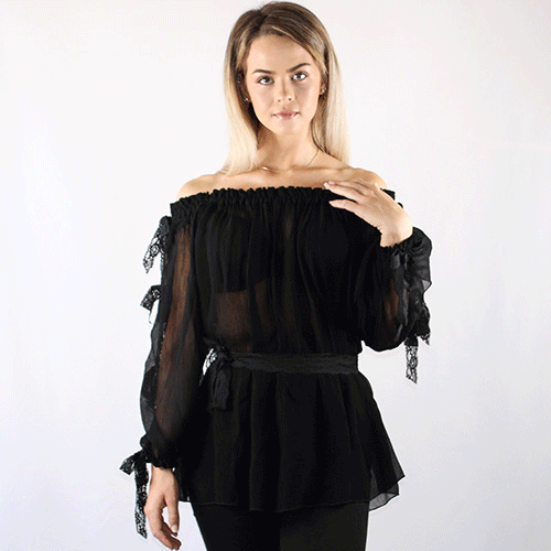 Woman with straight blonde hair wearing an off-the-shoulder, sheer black blouse with ruffled sleeves and lace details, standing against a plain white background.