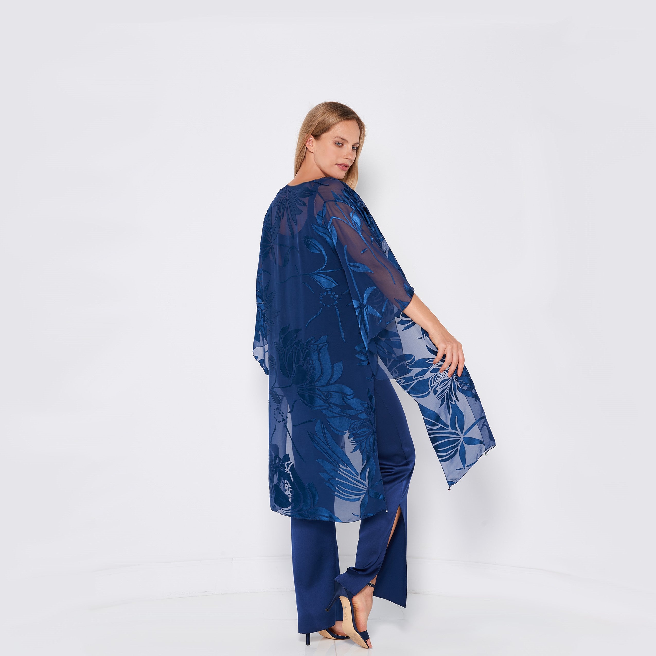 A woman wearing a sheer, blue, floral-patterned kimono over matching blue pants and high heels stands against a white background, looking over her shoulder and smiling slightly.