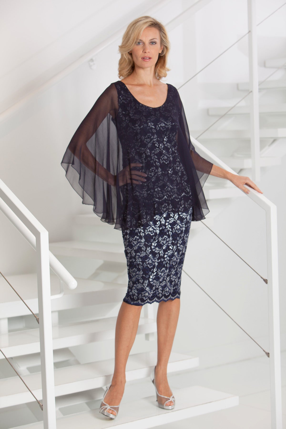 Woman wearing a dark blue silver lace dress with sheer overlay on a white staircase.