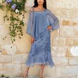 Woman in a blue dress standing against a stone wall with greenery.