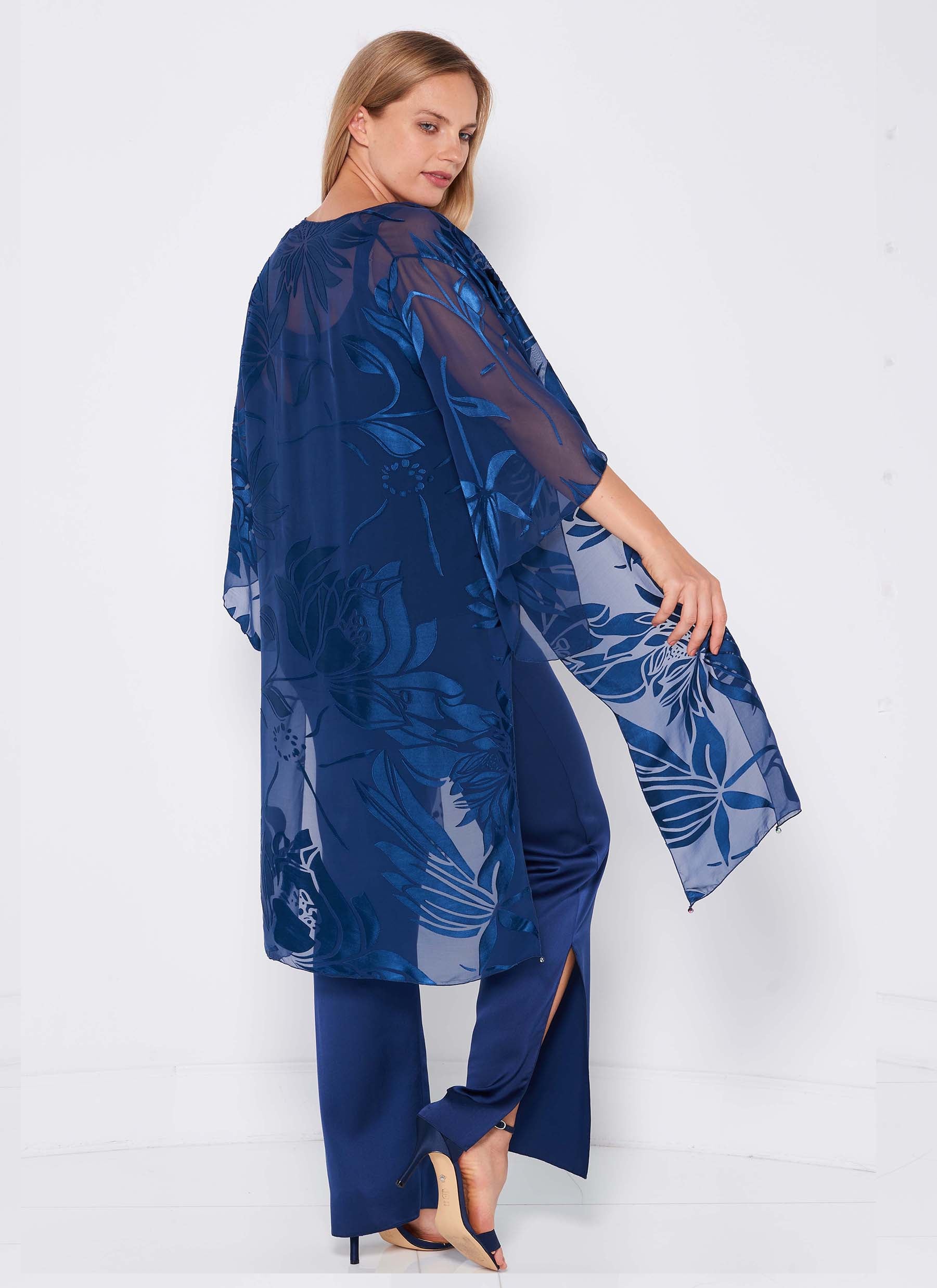 A woman models the Sara Mique Evening Wear Devore Duster Pant Set K1809H/Cami/P1703 in sheer indigo over matching pants and heels, standing sideways and looking over her shoulder against a white background.
