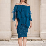 Woman wearing a blue off-shoulder blouse with ruffled layers and a skirt in front of a stone building.