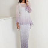 A woman wears the Sara Mique Evening Wear T1501OM/D1564SCLOM gown indoors—a long, gradient dress fading from white to lavender, with a sheer plissé top and flared sleeves. She finishes the look with silver open-toe heels.