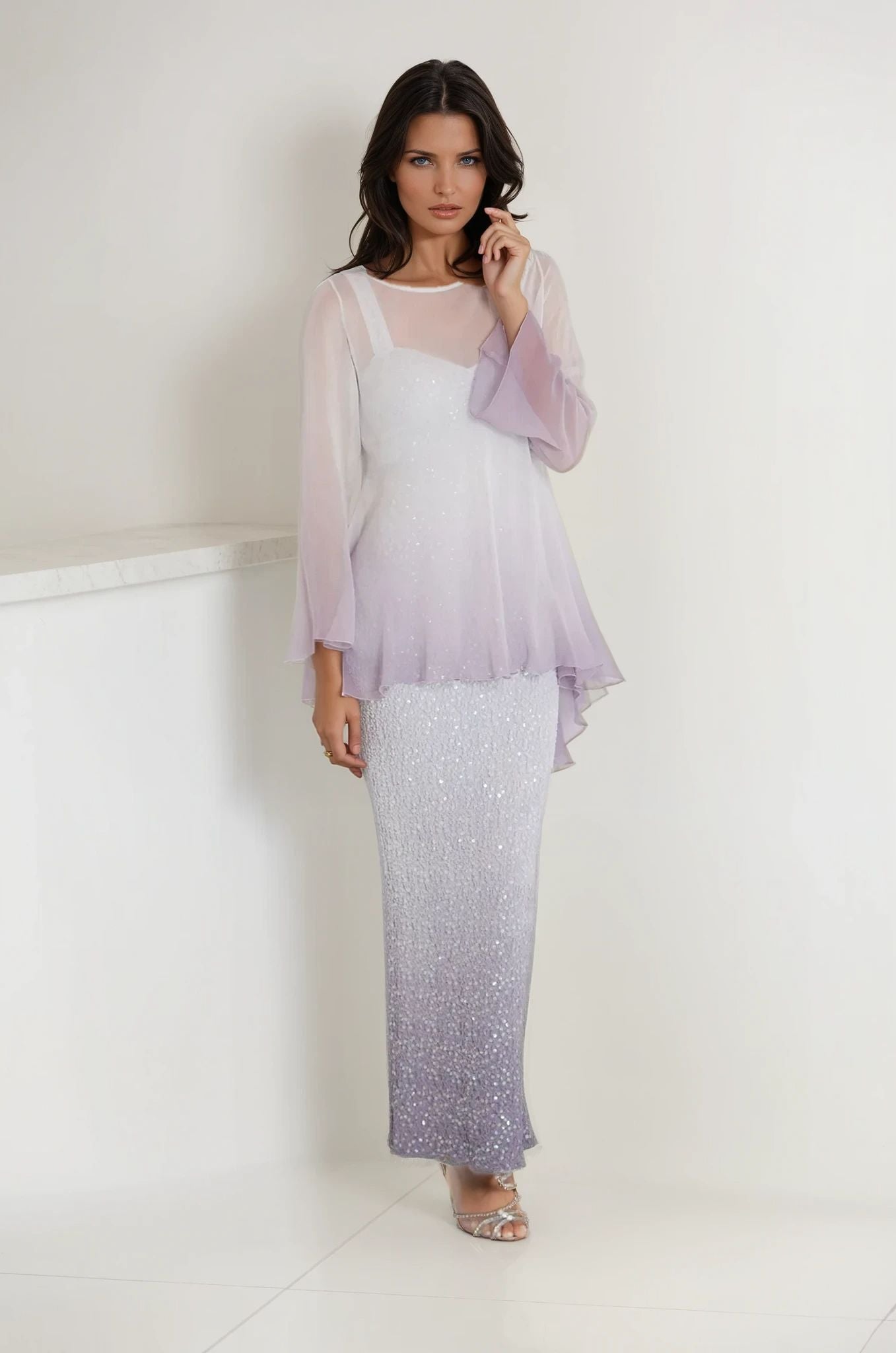A woman wears the Sara Mique Evening Wear T1501OM/D1564SCLOM gown indoors—a long, gradient dress fading from white to lavender, with a sheer plissé top and flared sleeves. She finishes the look with silver open-toe heels.